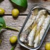 Imagem de uma lata de sardinha aberta com azeitonas em volta na mesa