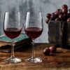 Foto mostra duas taças de vinho tinto em cima da mesa ao lado de um cacho de uvas.