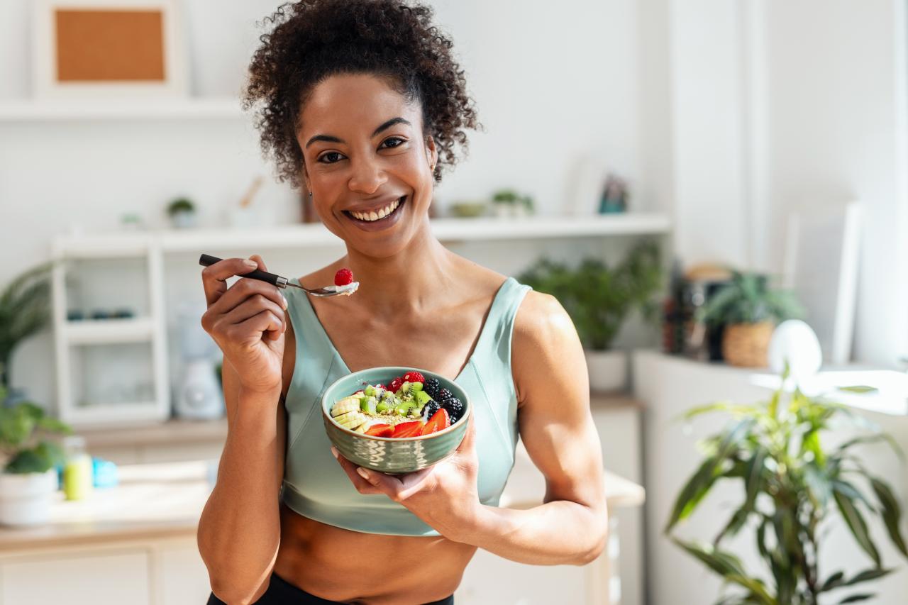Mulher negra sorrindo segurando uma cumbuca com frutas e legumes.