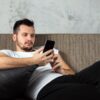 Homem sentado no sofá mexendo no celular.