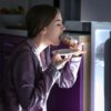 Imagem de uma mulher, a noite, em frente à geladeira, comendo um doce; ela veste um pijama roxo de mangas compridas