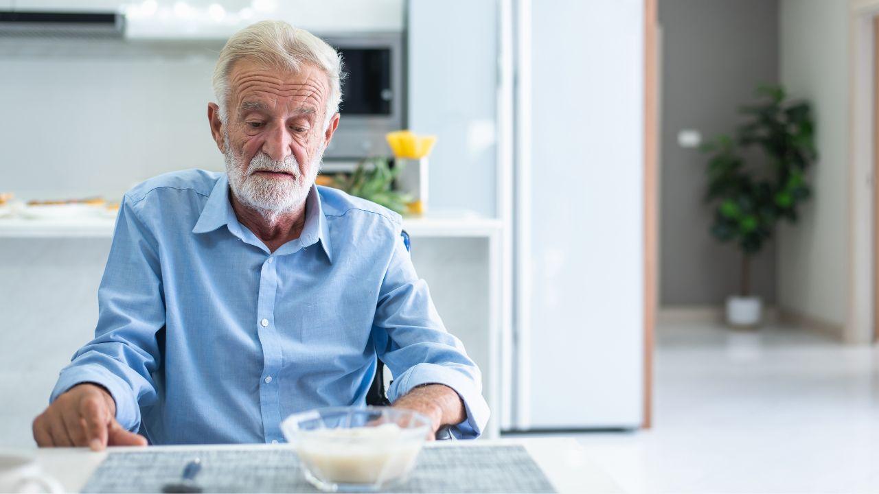Imagem de um idoso sentado à mesa olhando para um pote de vidro com algo branco dentro