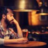 Foto mostra homem sentado em um bar bebendo uma taça de cerveja.