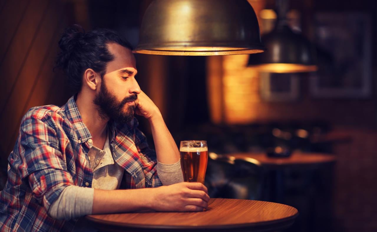Foto mostra homem sentado em um bar bebendo uma taça de cerveja.