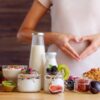 Foto mostra uma mulher fazendo coração com a mão na barriga. Na frente de uma mesa com comidas de fibras.
