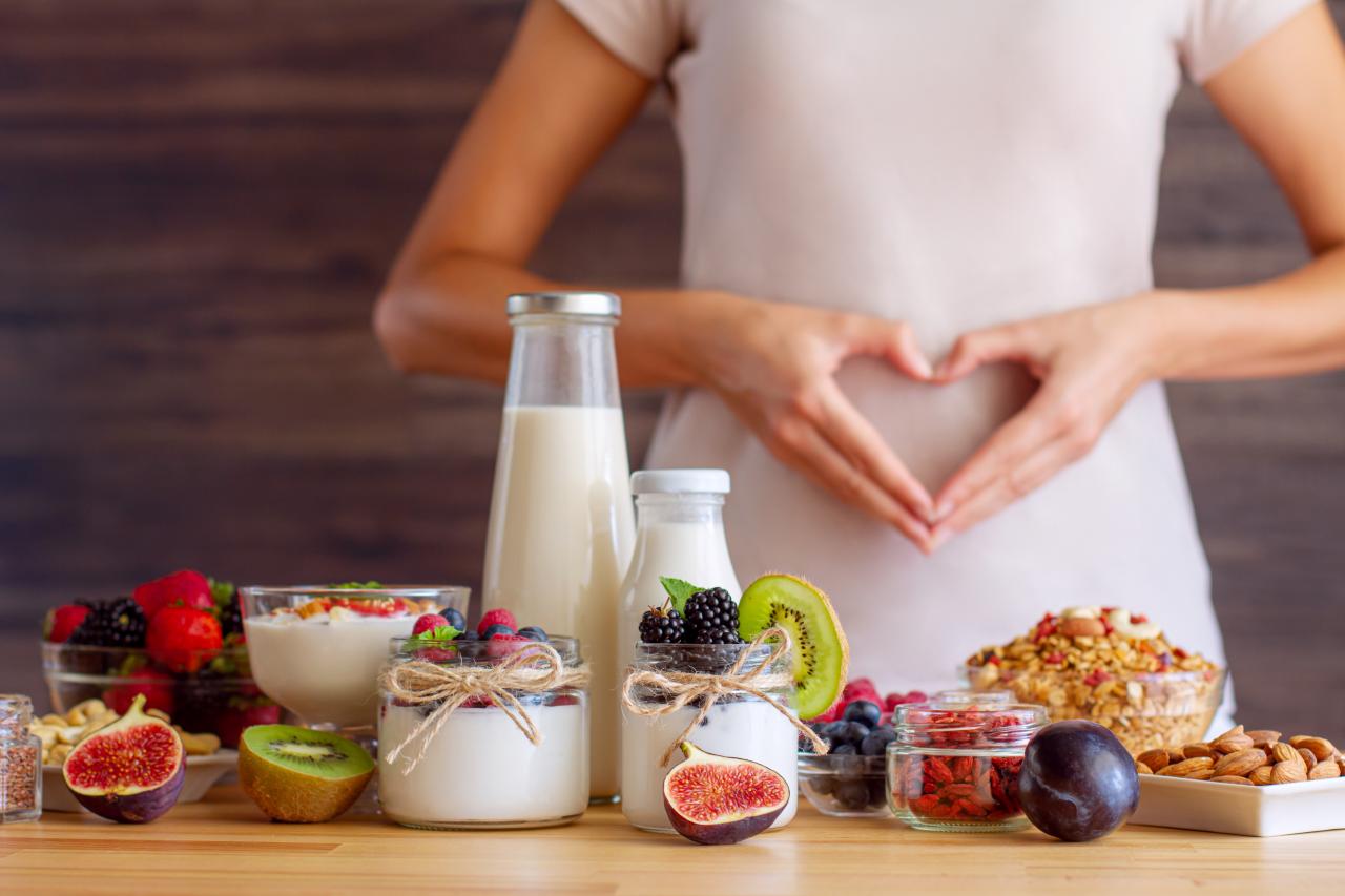 Foto mostra uma mulher fazendo coração com a mão na barriga. Na frente de uma mesa com comidas de fibras.