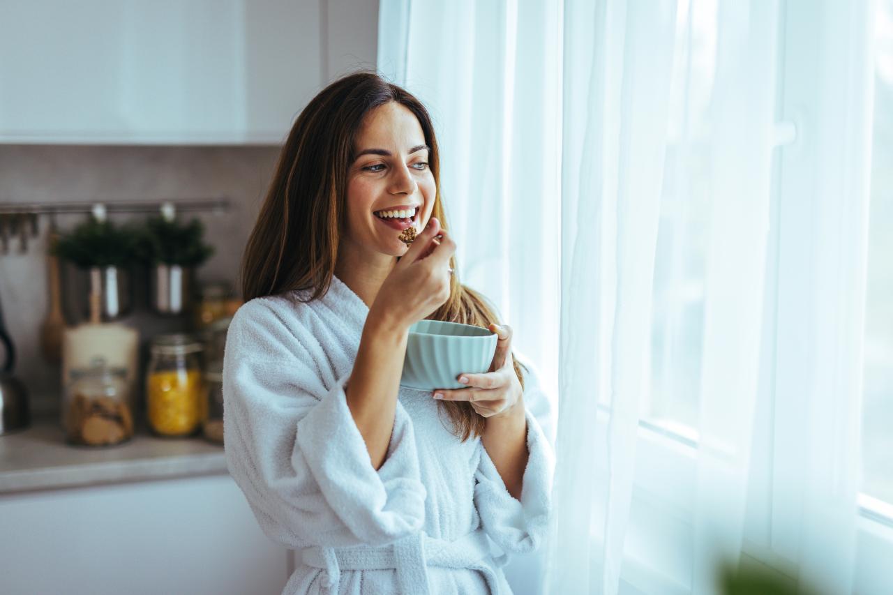 Mulher em pé na cozinha comendo um alimento na cumbuca com colher.
