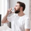 Homem branco, com barba e cabelo castanho sentado na cama, bebendo um copo de água.