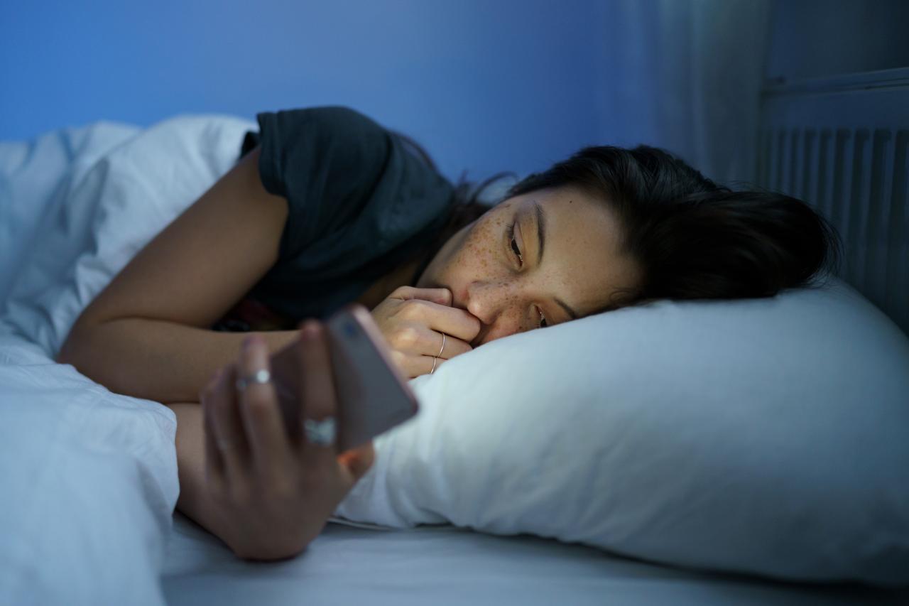Mulher deitada na cama no escuro mexendo no celular.