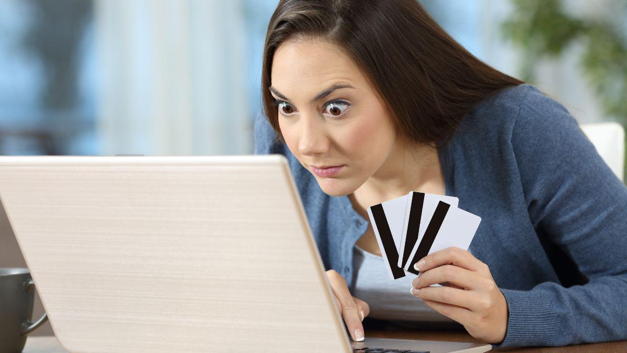 Imagem de uma mulher com os olhos saltados olhando para o computador e segurando três cartões de crédito