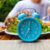 Imagem de um relógio azul em cima de uma mesa com pratos de comida atrás