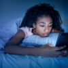 Mulher negra deitada na cama no escuro e mexendo no celular.