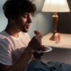 Homem no escuro comendo um pão sentado na cama.