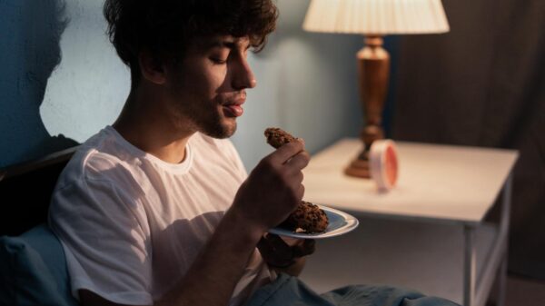 Homem no escuro comendo um pão sentado na cama.