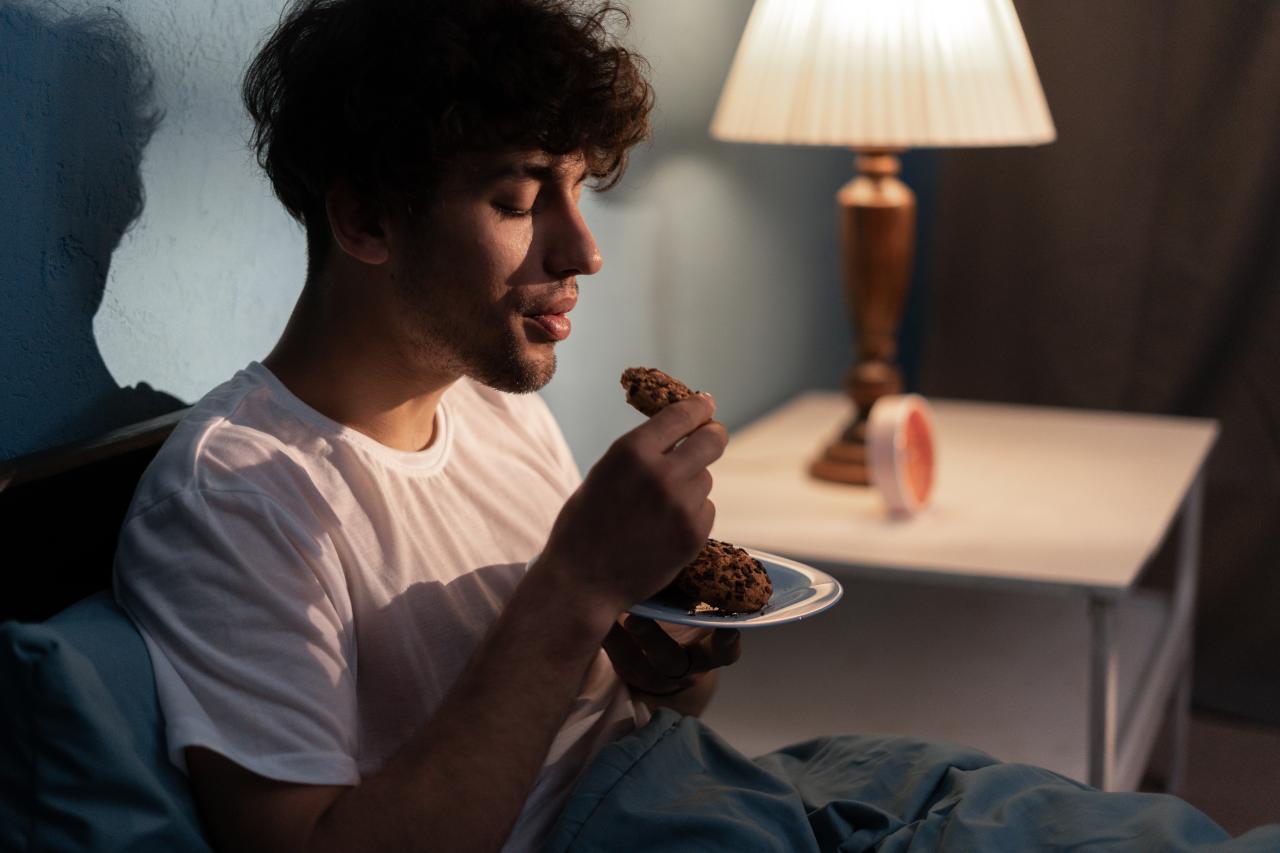 Homem no escuro comendo um pão sentado na cama.