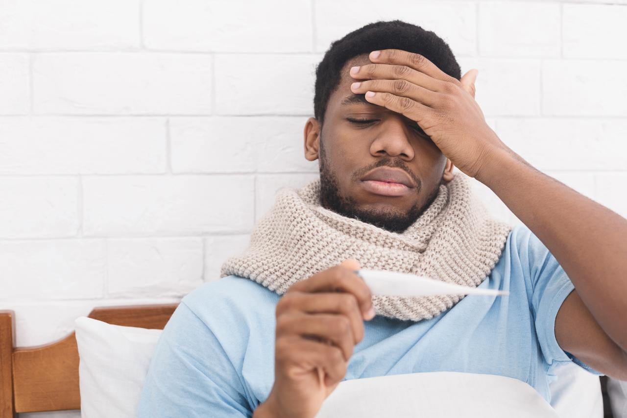 Homem sentado na cama com um cachecol, com a mão na testa, e segurando um termômetro.
