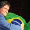 Foto mostra Roberto Carlos de terno azul abraçando uma pelúcia da bandeira do Brasil no palco.