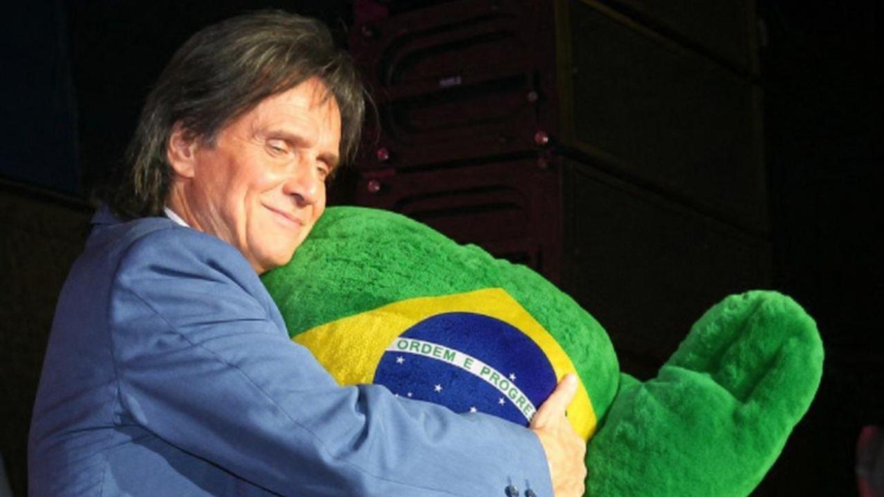 Foto mostra Roberto Carlos de terno azul abraçando uma pelúcia da bandeira do Brasil no palco.