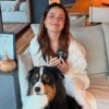 Juliette sentada no sofá e um cachorro preto e branco sentado no chão. Mulher branca, cabelo castanho médio, ela sorrindo.