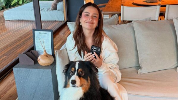Juliette sentada no sofá e um cachorro preto e branco sentado no chão. Mulher branca, cabelo castanho médio, ela sorrindo.