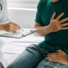 Foto mostra homem sentado no consultório com a mão no peito enquanto uma médica o consulta.