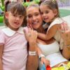 Foto mostra Virginia sentada na cadeira com as filhas ao lado.