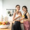 Duas mulheres na cozinha com frutas nas mãos e no cesto da bancada da cozinha.