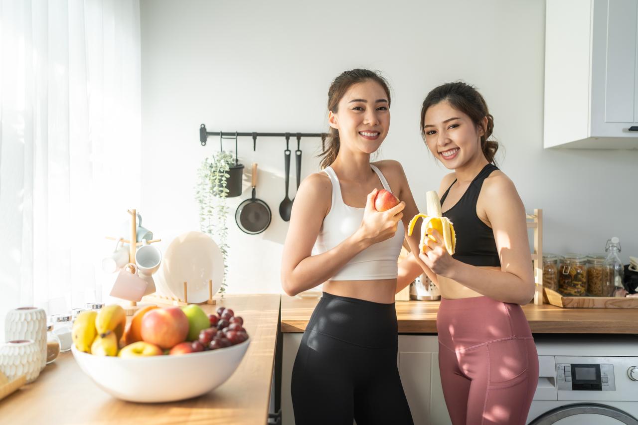 Duas mulheres na cozinha com frutas nas mãos e no cesto da bancada da cozinha.