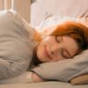 Foto mostra mulher deitada na cama dormindo com lençóis brancos.