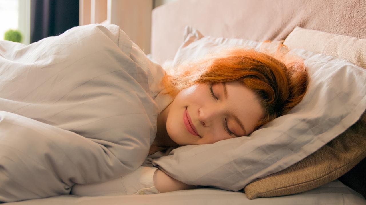Foto mostra mulher deitada na cama dormindo com lençóis brancos.