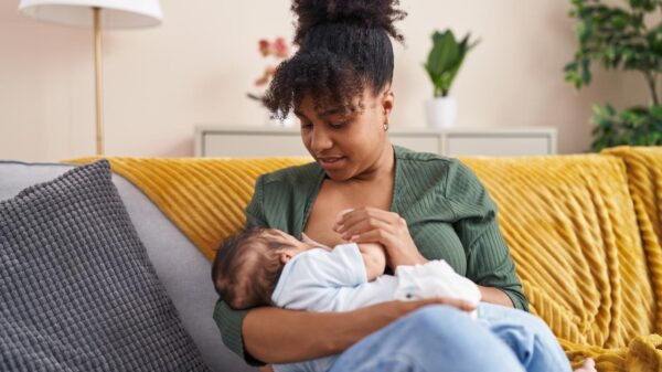 Mulher negra sentada no sofá, cabelo, amamentando bebê no colo.