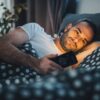 Homem deitado na cama com o celular na mão com a tela brilhando o rosto dele.