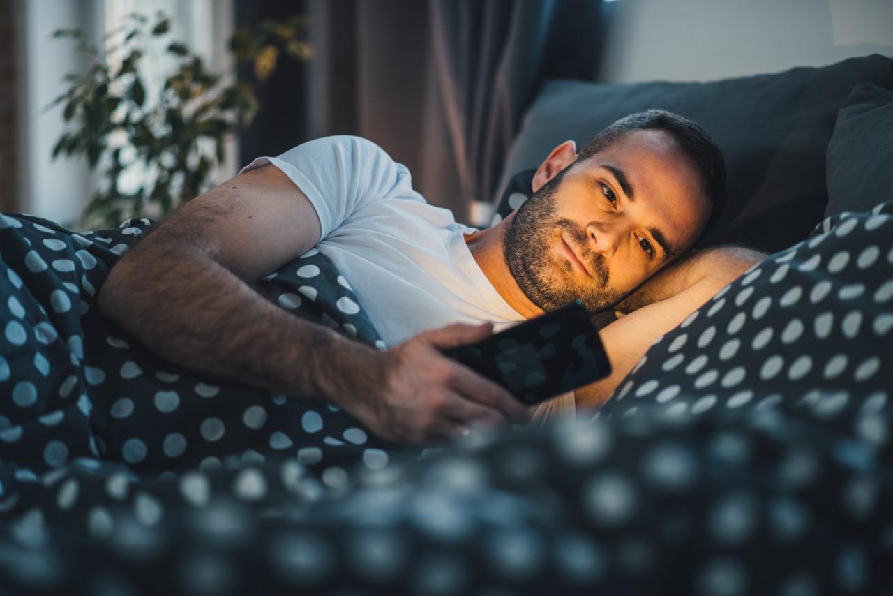 Homem deitado na cama com o celular na mão com a tela brilhando o rosto dele.