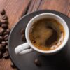 Foto mostra uma xícara com café cheia em cima de uma mesa de madeira com grãos de café ao lado.