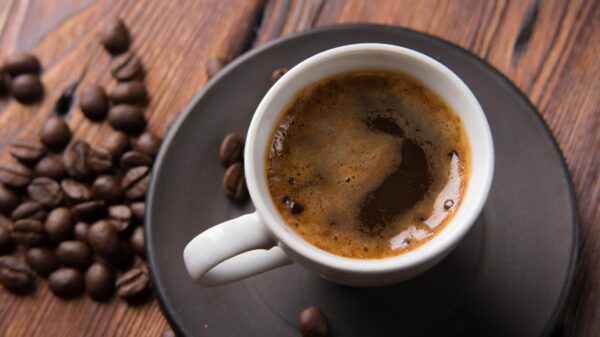 Foto mostra uma xícara com café cheia em cima de uma mesa de madeira com grãos de café ao lado.