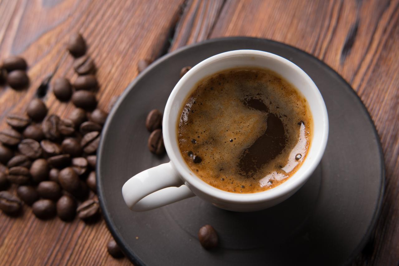 Foto mostra uma xícara com café cheia em cima de uma mesa de madeira com grãos de café ao lado.