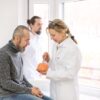 Foto mostra um homem em um consultório médico com uma médica cuidando dele.