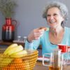 Mulher idosa com cabelo branco sentada em uma mesa com frutas e sorrindo.
