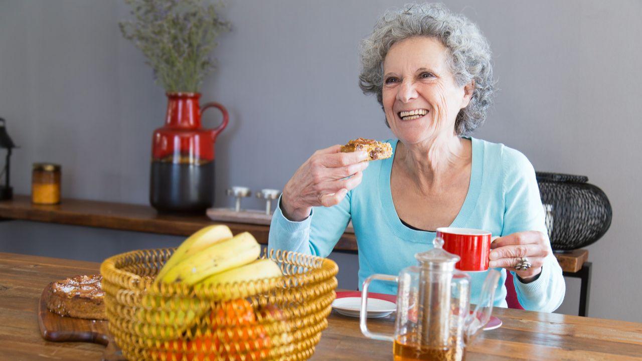 Mulher idosa com cabelo branco sentada em uma mesa com frutas e sorrindo.