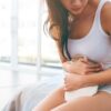 Mulher jovem sentada na cama com o abdômen encurvado e com uma bolsa térmica na barriga.