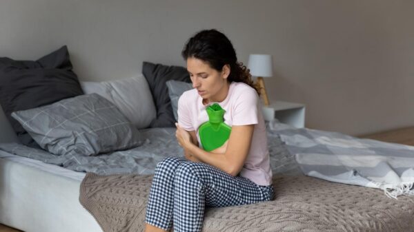 Foto mostra mulher com dor sentada na cama segurando uma bolsa térmica.