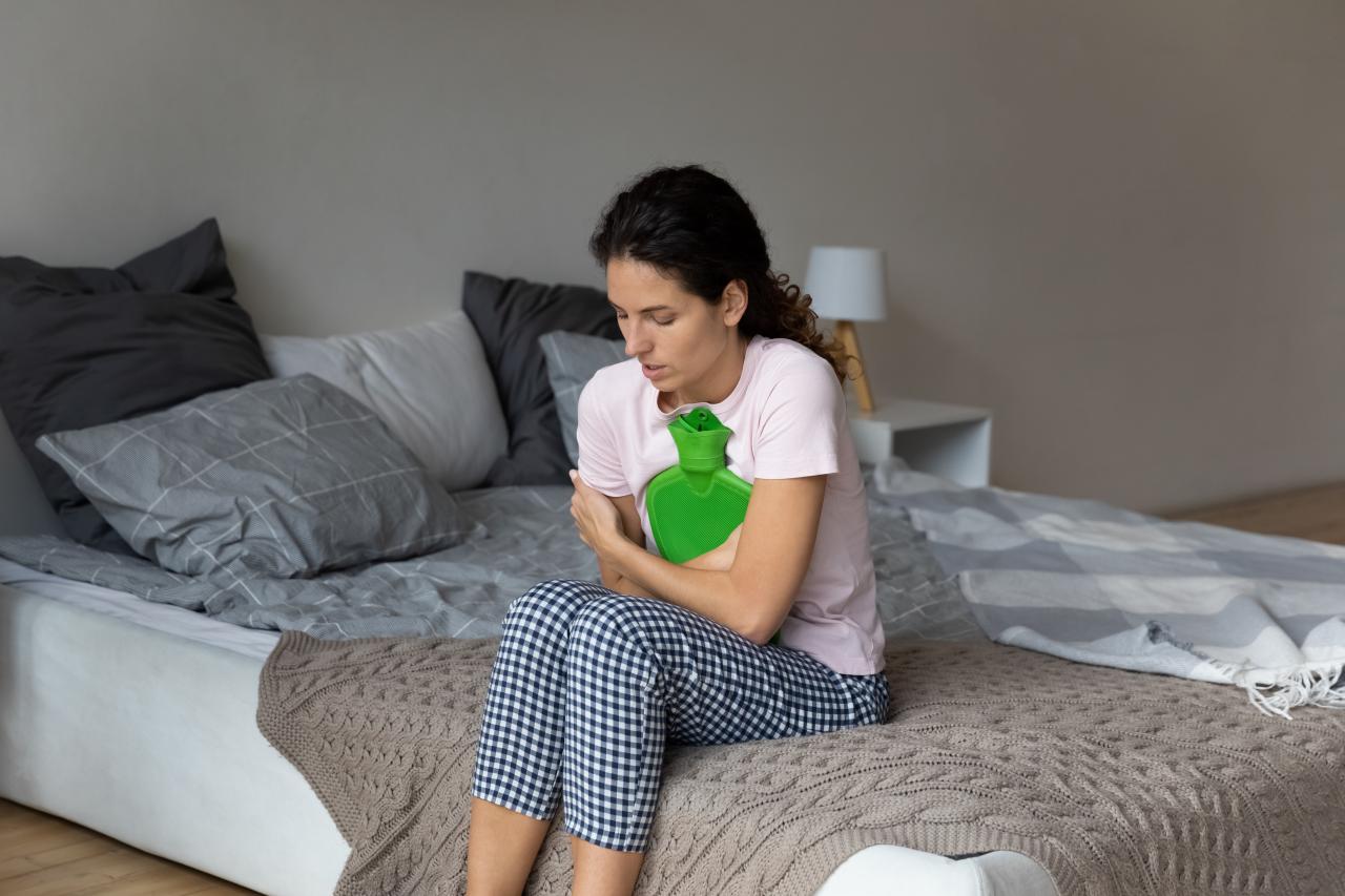 Foto mostra mulher com dor sentada na cama segurando uma bolsa térmica.