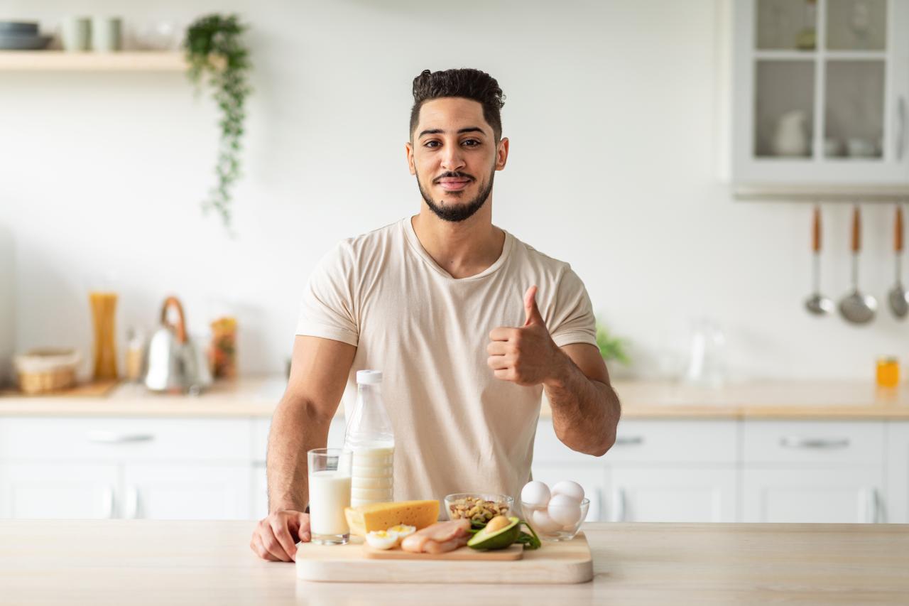Homem em pé na cozinha com leite em garrafa e outras comidas em cima da bancada da cozinha.