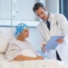 Foto mostra uma mulher na cama de hospital com lenço na cabeça, e um médico explicando coisas para ela na prancheta.