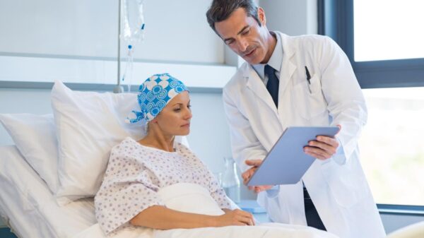 Foto mostra uma mulher na cama de hospital com lenço na cabeça, e um médico explicando coisas para ela na prancheta.