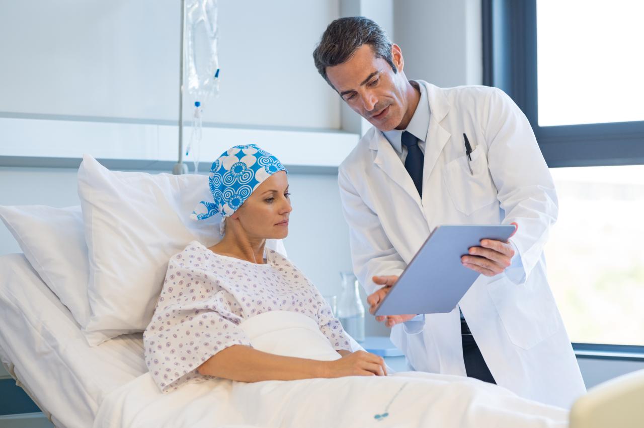 Foto mostra uma mulher na cama de hospital com lenço na cabeça, e um médico explicando coisas para ela na prancheta.